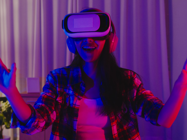 A woman wearing a VR headset and headphones in a room lit pink and blue, smiling with both hands raised as she explores a virtual space.